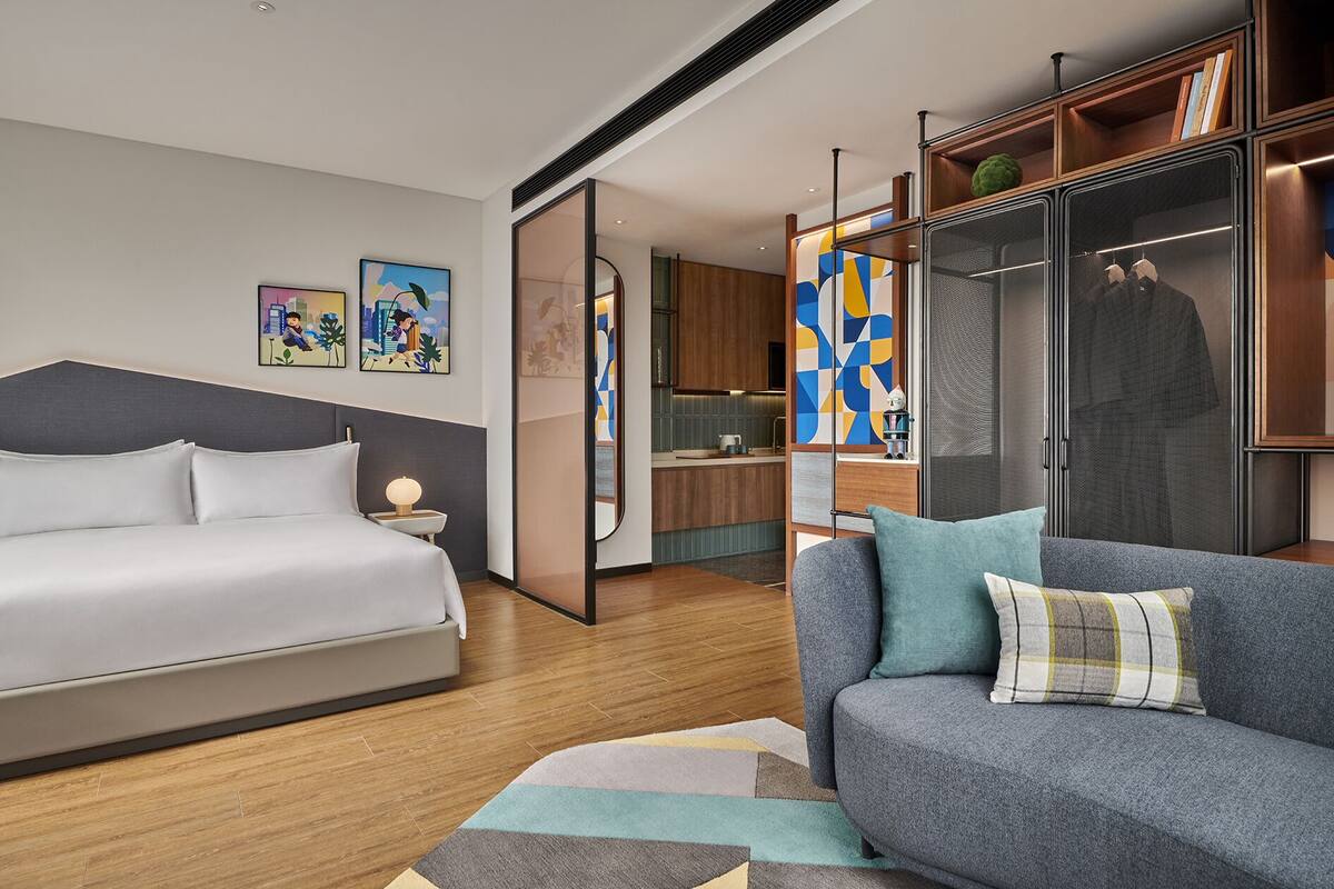 Une chambre d’hôtel moderne, équipée d’un lit, d’un canapé, d’une petite table avec une lampe, et d’un coin cuisine visible à travers une porte ouverte.