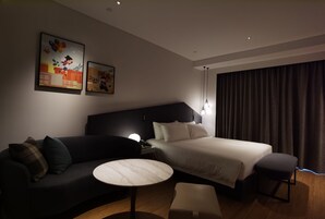 A modern hotel room with a dark-colored sofa, a round table, a bed with white linens, and two wall-mounted artworks.