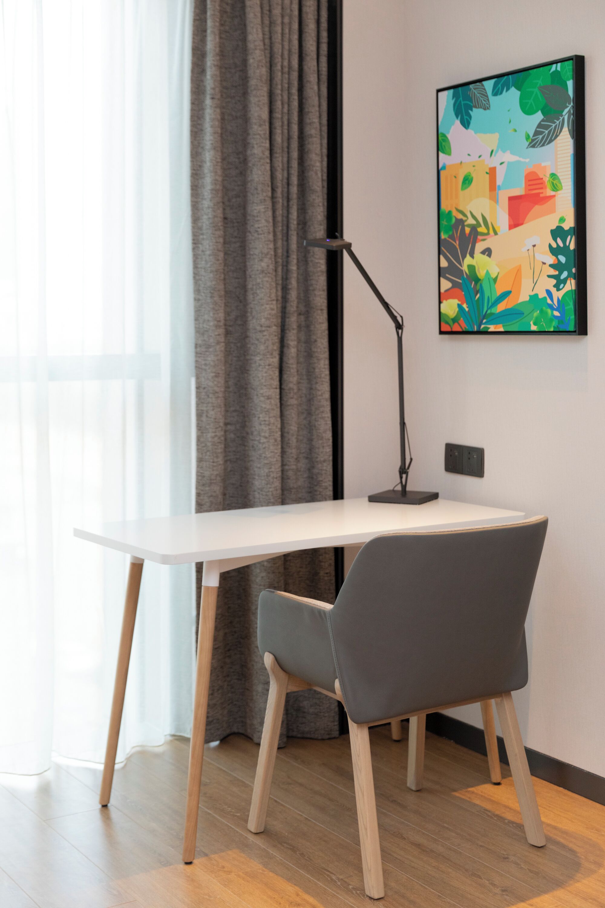 Un espacio de oficina moderno con un escritorio blanco, una silla gris, una lámpara de pie y un cuadro abstracto colorido en la pared.