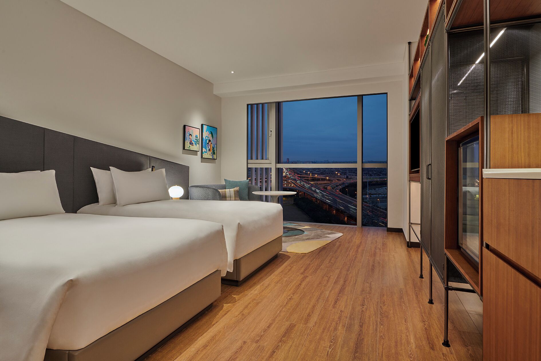 Ein Hotelzimmer mit zwei Betten, einem großen Fenster mit Blick auf die Stadt und einem kleinen Tisch mit einer Lampe.