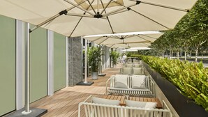 Un espacio de estar al aire libre con sillas y mesas de metal blanco, cubierto por grandes sombrillas color beige, y delimitado por un seto verde.