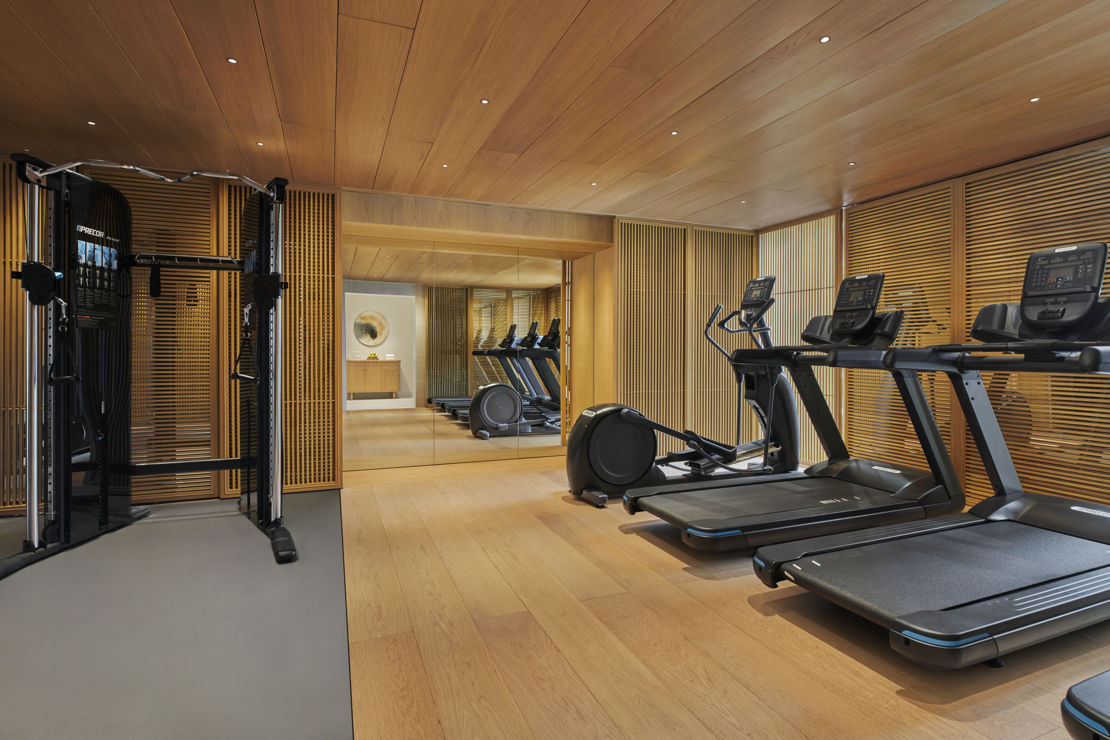 A modern gym with wooden floors and walls, equipped with treadmills, elliptical machines, and weightlifting stations.