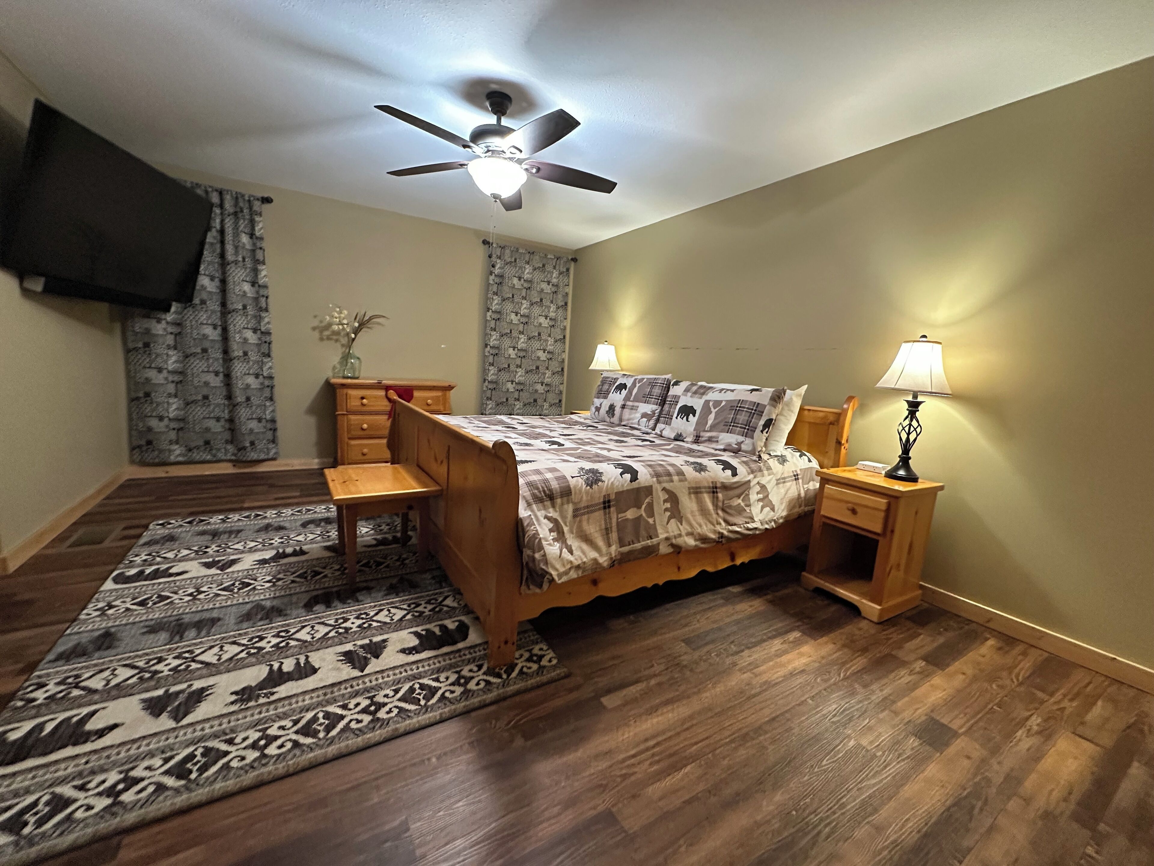 A bedroom with a wooden bed, two bedside tables with lamps, a ceiling fan, a patterned rug, and curtains.