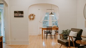 Un salon spacieux doté d’un plancher en bois, un coin repas avec une table et des chaises, et une grande fenêtre agrémentée de rideaux.