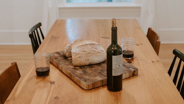 Une table de salle à manger en bois, sur laquelle on trouve un pain, une bouteille de vin et un verre de vin, le tout posé sur une planche à découper.