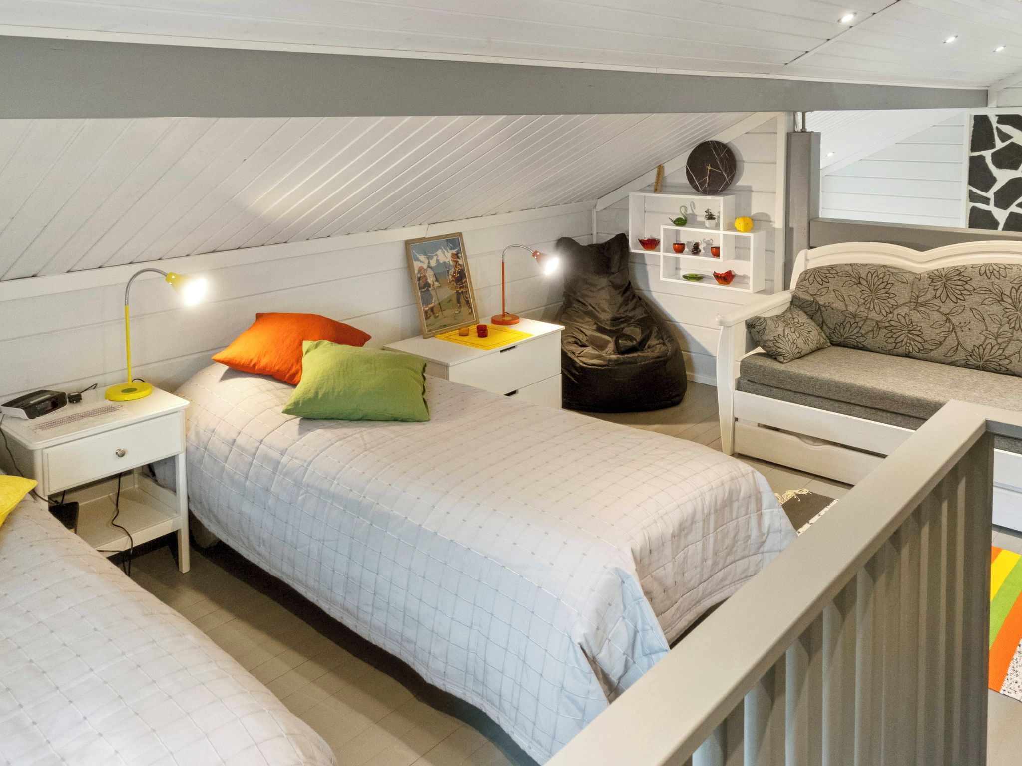Un dormitorio con cama, mesitas de noche, un escritorio, una silla, un sillón de felpa y un estante con objetos decorativos.