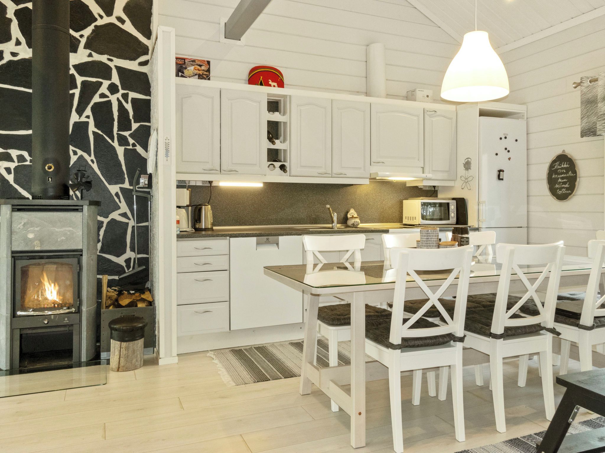 Una cocina moderna con armarios blancos, una pared de piedra, estufa de leña y un comedor con sillas blancas y una mesa de vidrio.