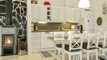 Una cocina moderna con armarios blancos, una pared de piedra, estufa de leña y un comedor con sillas blancas y una mesa de vidrio.