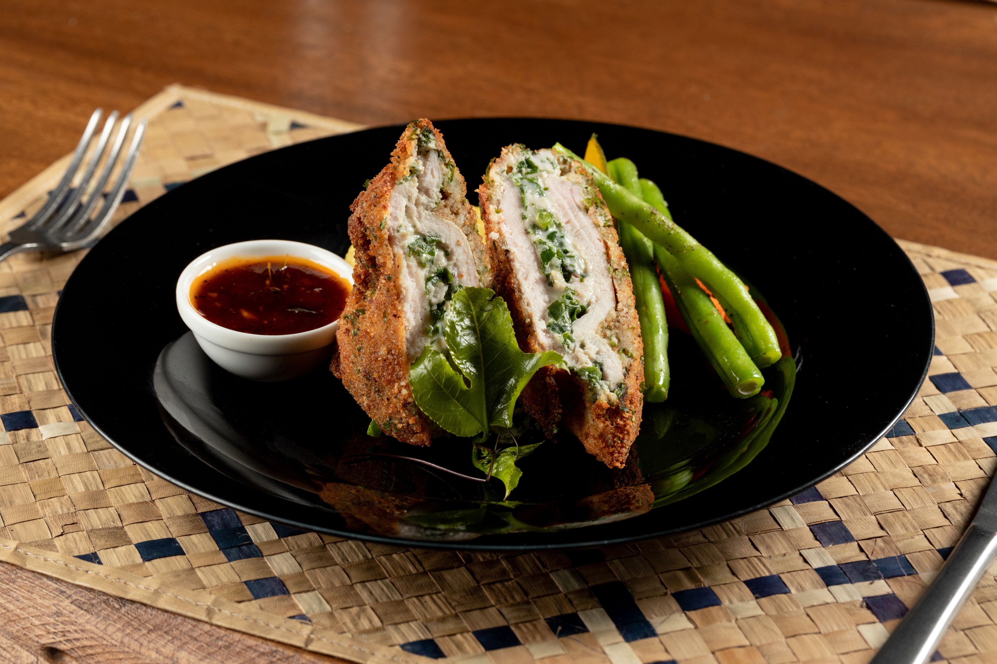 A plate with a breaded and sliced meat dish, green vegetables, and a small bowl of sauce on a woven placemat.