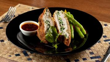 A plate with a breaded and sliced meat dish, green vegetables, and a small bowl of sauce on a woven placemat.