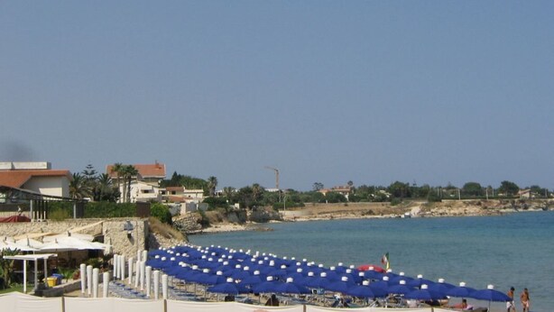 A beachside area with rows of blue umbrellas and beach chairs, a clear sky, and buildings in the background. - Appartamento pt in Villa Falari - Clima - Wi-fi (Avola)