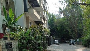 A residential street with a sidewalk, parked cars, and a building with a stone wall and greenery. - Room-nice Boutique Guest House In South (New Delhi)