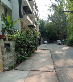 A residential street with a sidewalk, parked cars, and a building with a stone wall and greenery. - Room-nice Boutique Guest House In South (New Delhi)