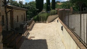 Un chemin de gravier mène à un bâtiment en pierre, doté d’une rambarde en métal et d’une clôture.