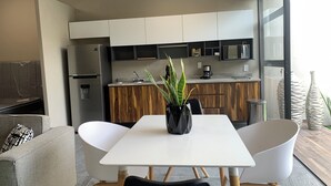 Een moderne keuken met witte kastjes, een inbouwoven en een koffieapparaat. Er is een eethoek met een witte tafel en stoelen, en een bloempot op de tafel.