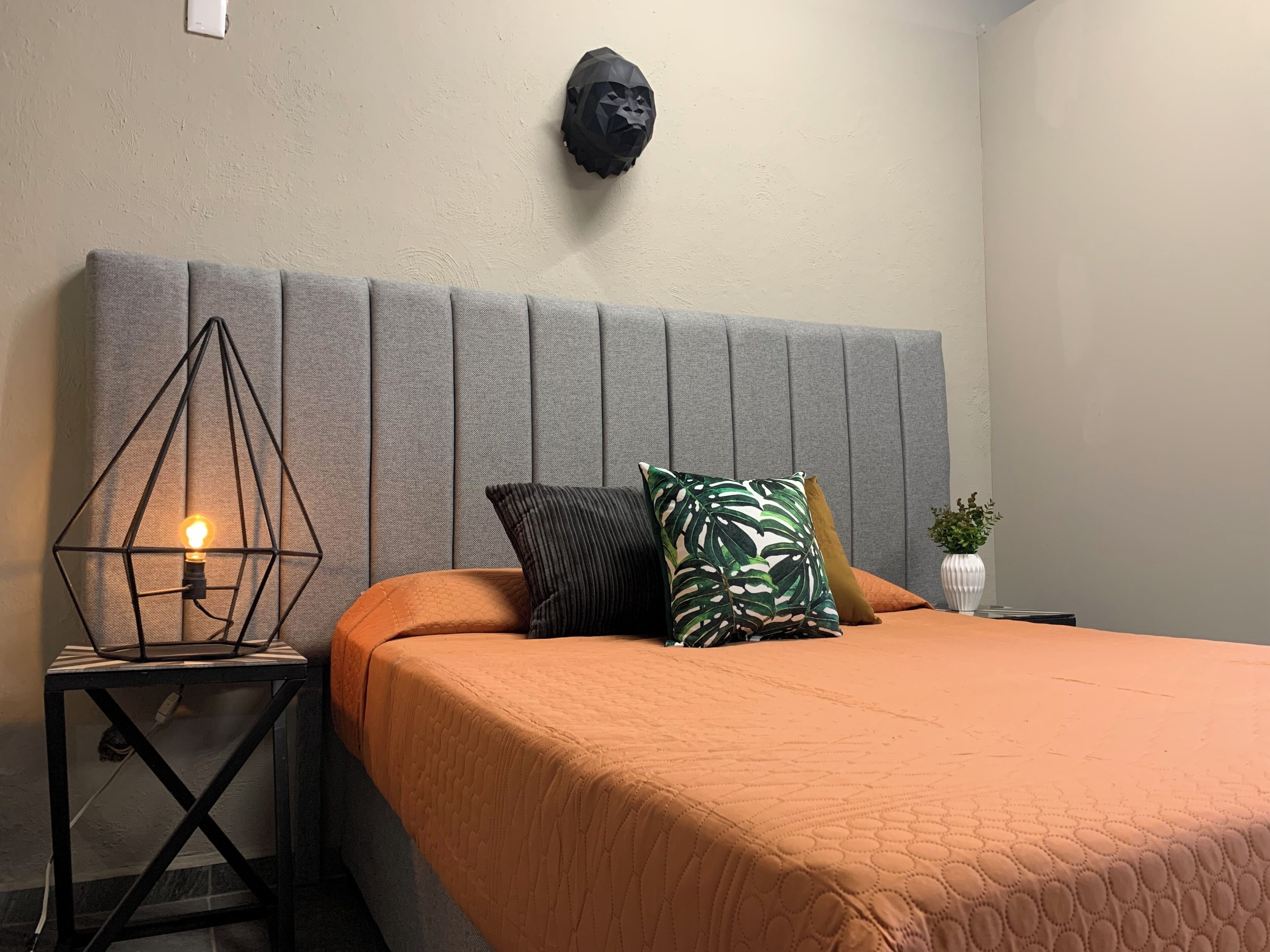 A bedroom with a grey upholstered headboard, a bed with an orange quilt, a nightstand with a geometric lamp, and a wall-mounted plant.