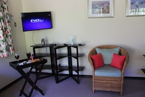 A room with a television displaying 'DSTV,' a wicker chair with red cushions, a small table with a tray containing a glass of wine, and a framed picture on the wall.