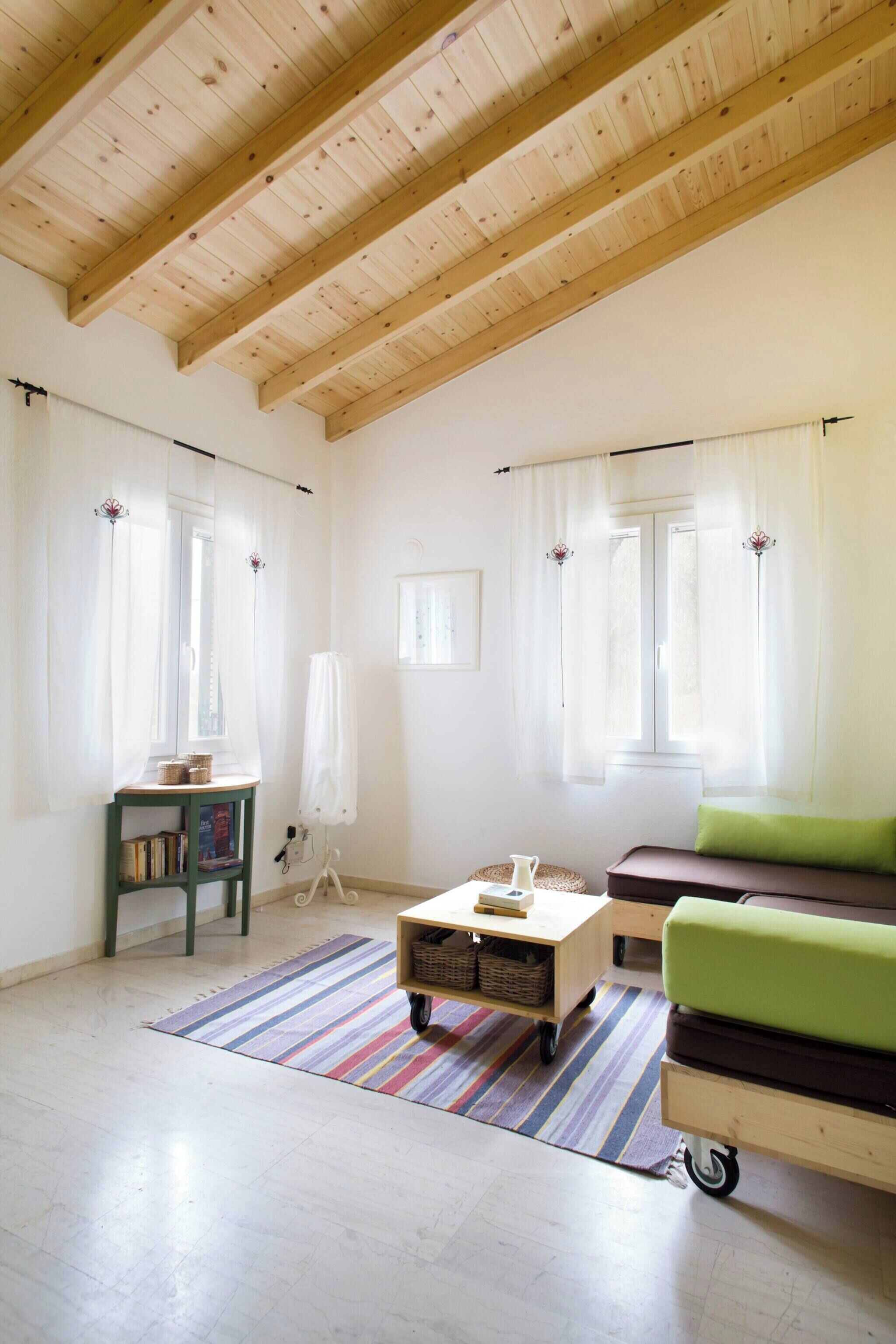 A room with a wooden ceiling, a green sofa, a small table, a standing lamp, and a window with curtains.