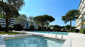 A rectangular swimming pool with clear water, surrounded by a tiled deck, trees, and modern buildings in the background. - Cozy Flat With Shared Pool (Caorle)