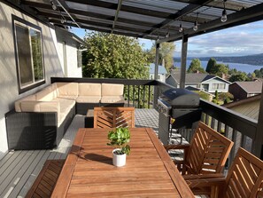Een overdekt terras buiten met een houten tafel, stoelen, een bank en een barbecue.