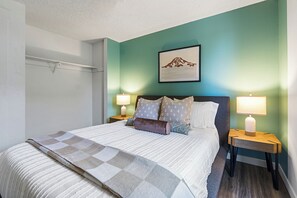Un lit bien fait, recouvert d’une couverture à carreaux, avec deux lampes de chevet et un tableau encadré accroché au mur.