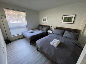 Een hotelkamer met twee bedden, een raam met zonwering, een nachtkastje met een lamp, en twee ingelijste foto’s aan de muur.