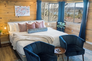 A bedroom with a bed, two blue chairs, a bedside table, a nightstand, a window with curtains, and a wall-mounted artwork.