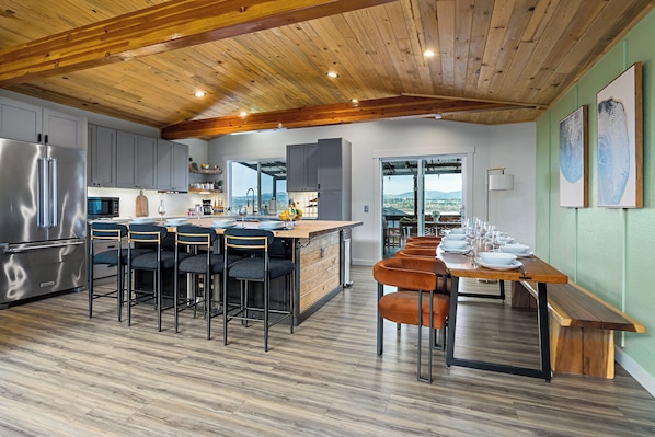 A modern kitchen with a wooden ceiling, grey cabinets, a central island, and a dining area with orange chairs.