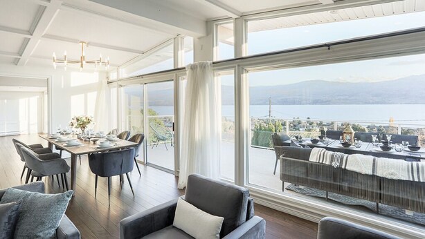 Ein geräumiger Essbereich mit Tisch für vier Personen, große Fenster mit Blick auf einen See und Berge, und ein Balkon mit Sitzgelegenheit im Freien.