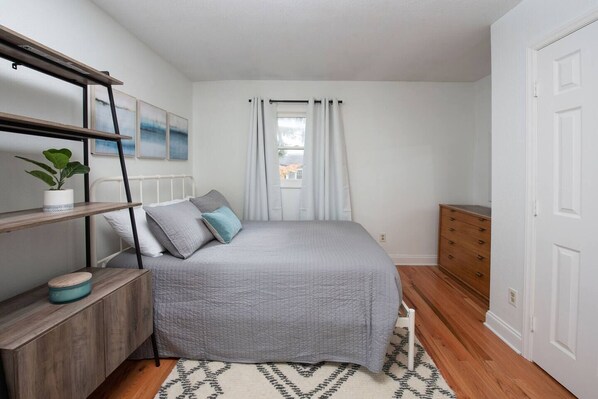 Un dormitorio con cama, mesita de noche, cómoda, ventana con cortinas y estante en la pared con una planta.