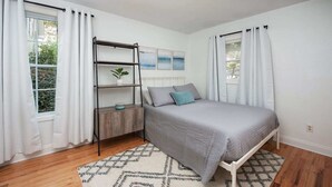 A bedroom with a bed, a wooden nightstand, a metal shelf, and a window with white curtains.