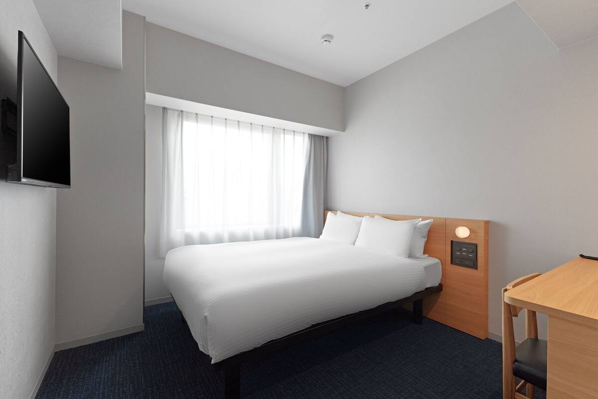 A hotel room with a bed, a TV mounted on the wall, a wooden desk, and a window with curtains.