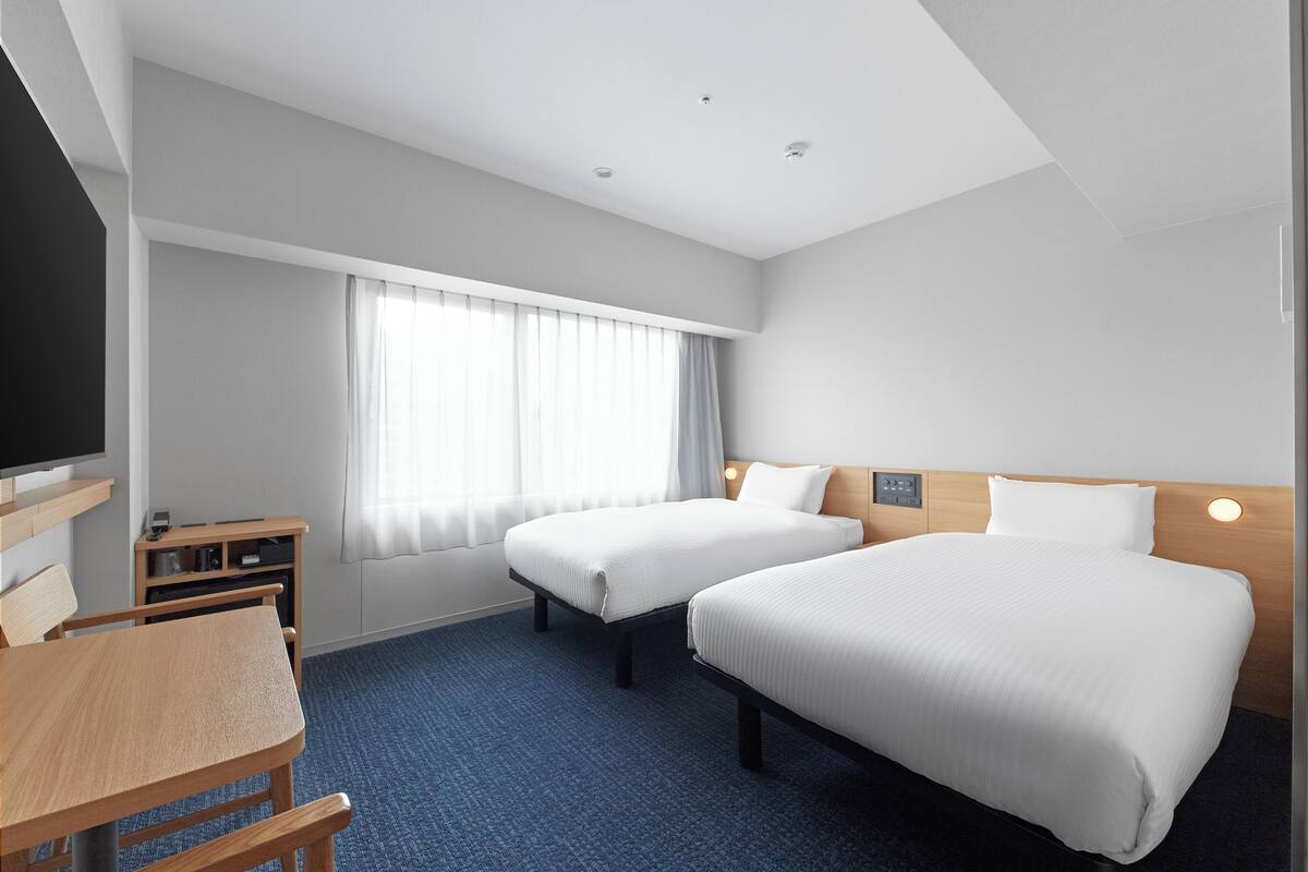 A hotel room with two beds, a desk, a chair, a TV, and a window with curtains.