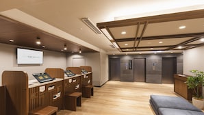 A modern hotel lobby with wooden reception desks, a large screen displaying the hotel name, and a seating area with a potted plant.