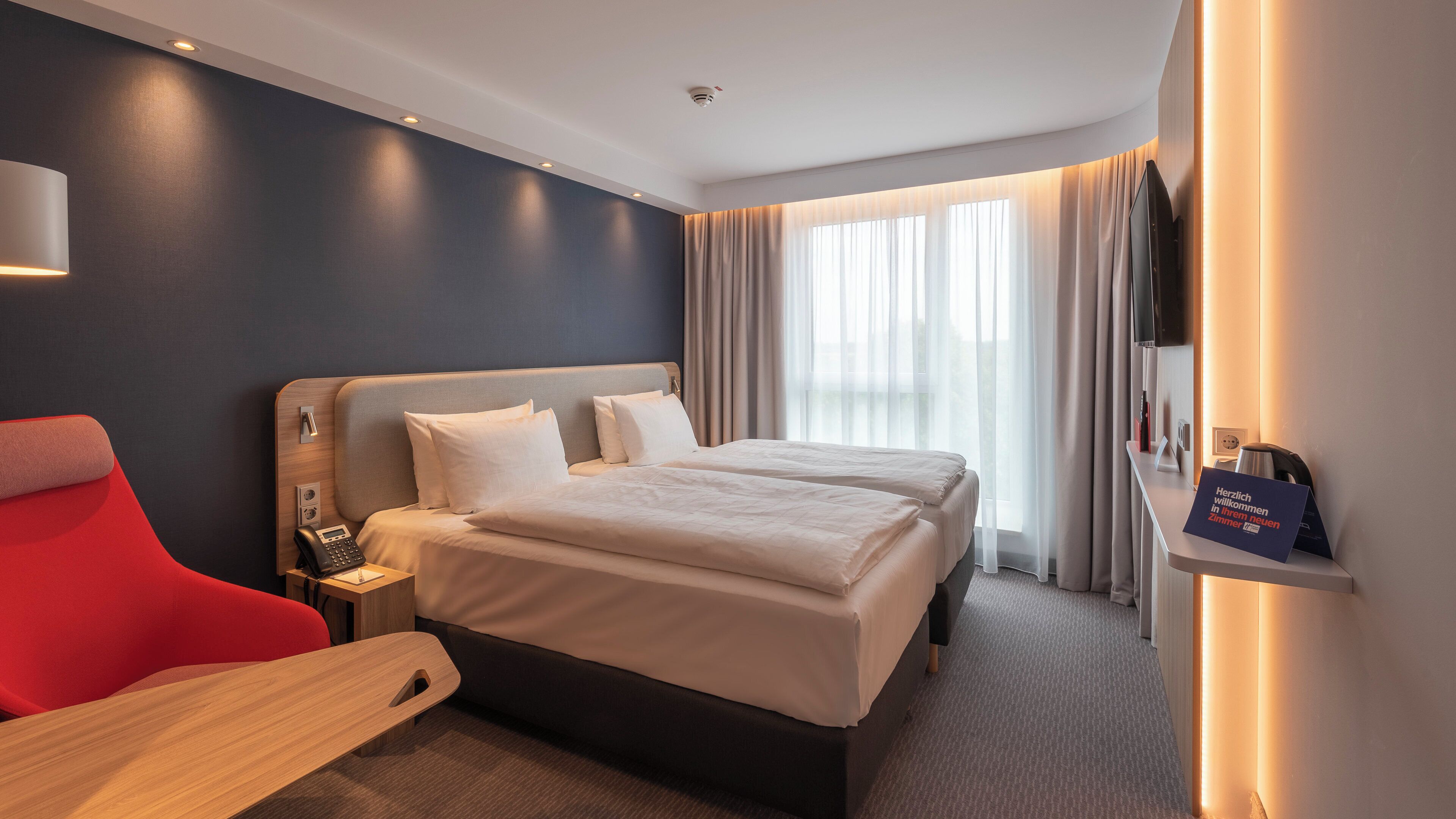 A hotel room with a bed, a desk, a chair, a TV, and a sign.