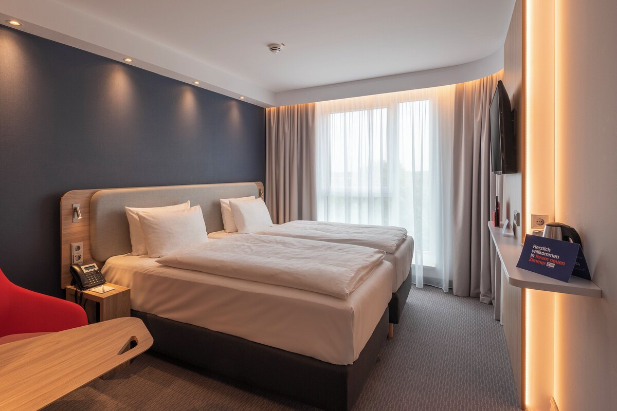 A hotel room with a bed, a desk, a chair, a TV, and a sign.