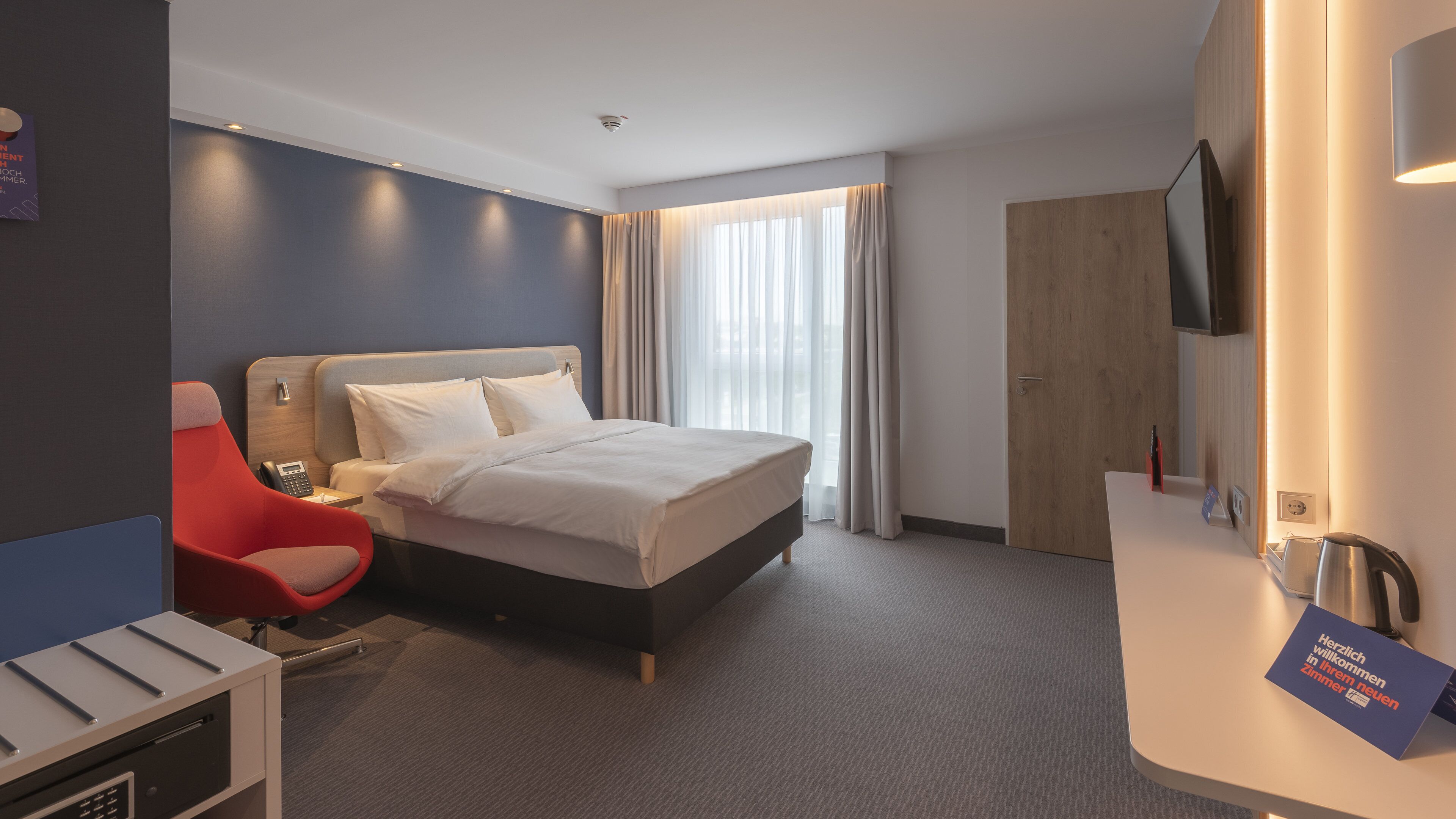 Una habitación de hotel con una cama, una silla roja, un escritorio con un teléfono, un televisor y un cartel en alemán.