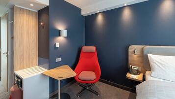 Habitación de hotel con una silla roja, una mesita, una mesita de noche con un teléfono y una cama con un almohadón.