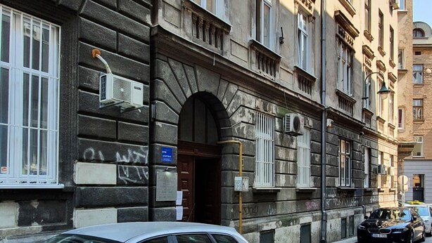 Une vue de rue avec une voiture blanche garée sur le bord, un bâtiment à l’architecture classique et un potelet d’incendie jaune.