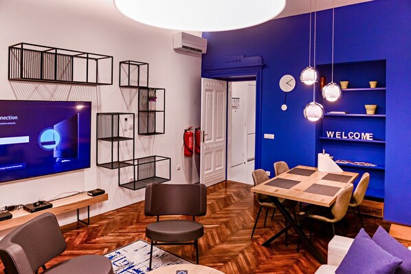 Una habitación moderna con piso de madera tipo espiga, un televisor mostrando una interfaz de red, un cartel de bienvenida y una pared azul con estantes.