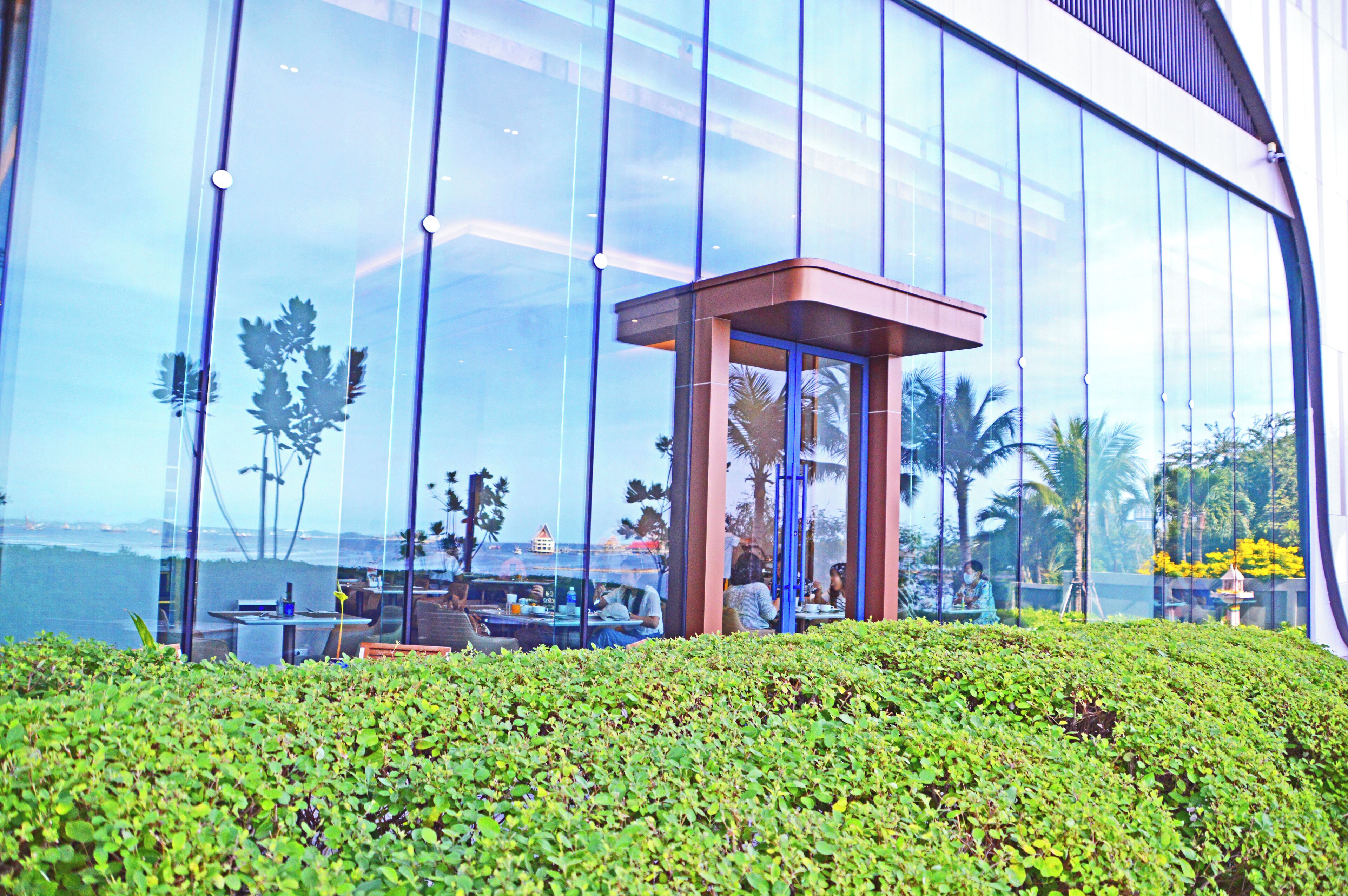 A modern building with large glass windows, a small enclosed area with a roof, and a well-maintained green hedge in the foreground.