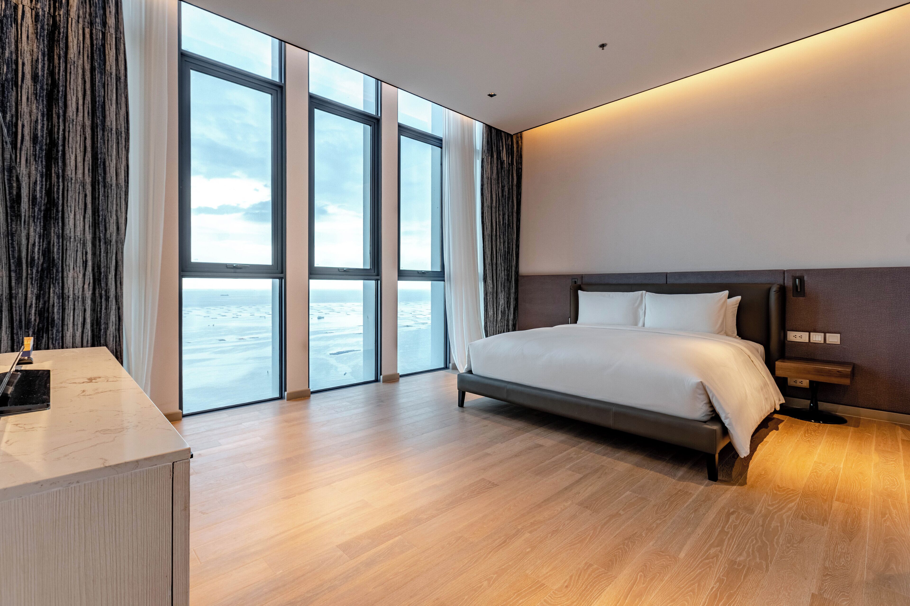 A modern hotel room with a large bed, a desk, and a view of the sea through floor-to-ceiling windows.