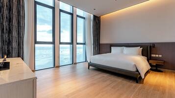 Una habitación de hotel moderna con una cama grande, un escritorio y vistas al mar a través de ventanas de gran altura.