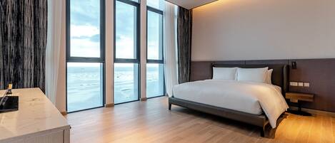 A modern hotel room with a large bed, a desk, and a view of the sea through floor-to-ceiling windows.