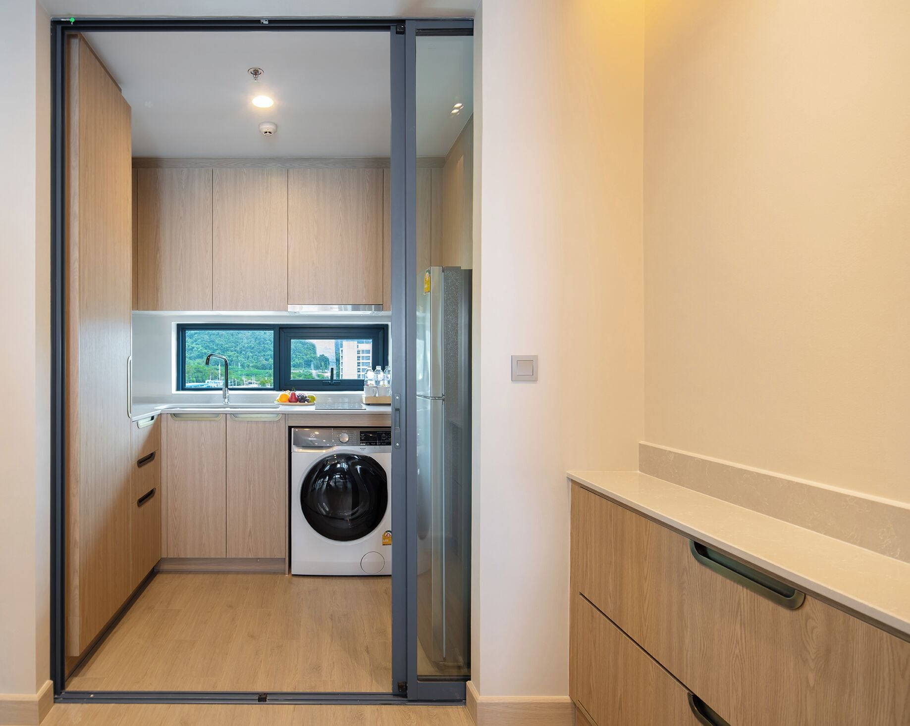 A compact kitchen with a washing machine, a refrigerator, and a window with a view of greenery.