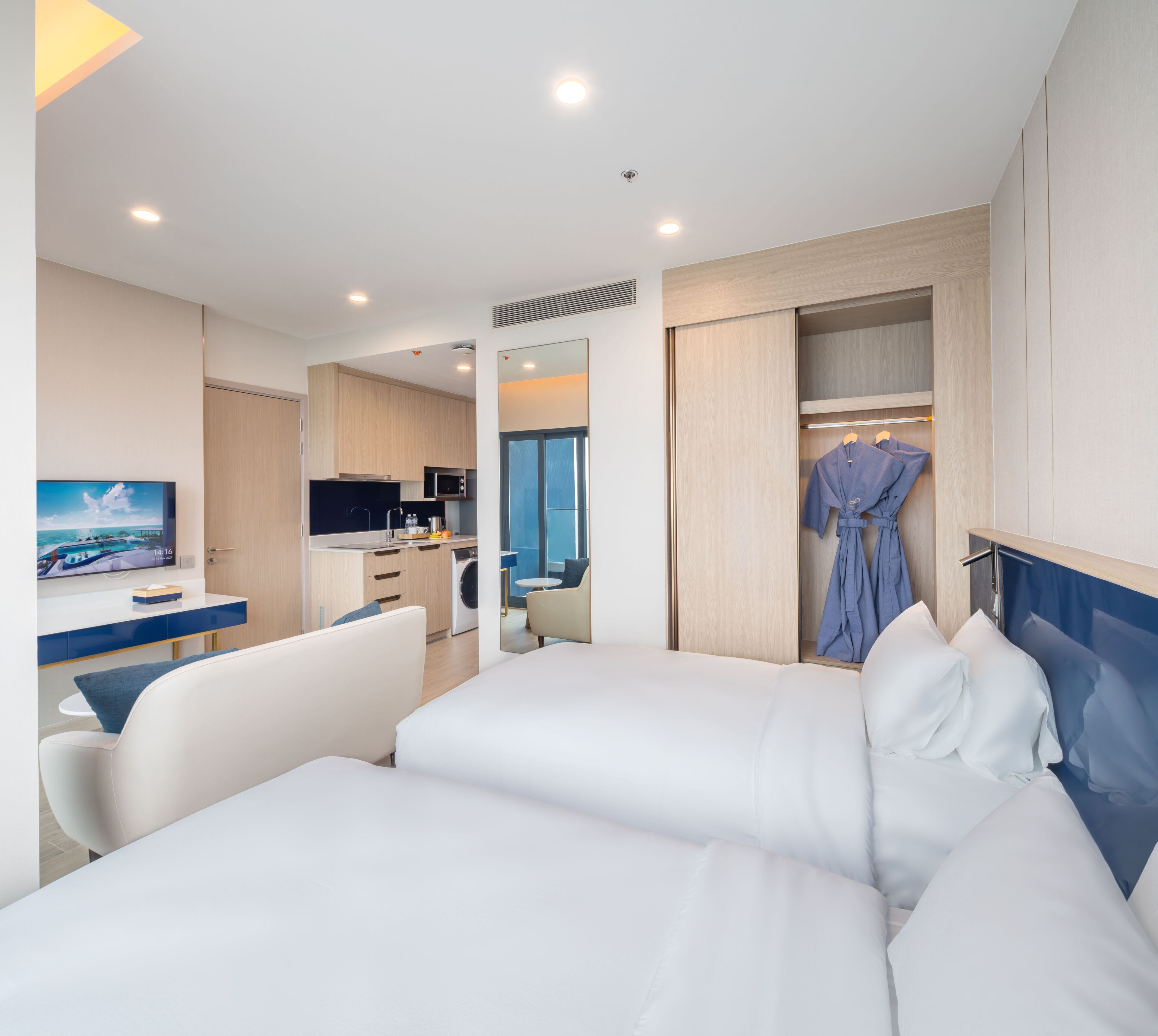Une chambre d’hôtel avec deux lits, un bureau avec une télévision, une kitchenette et une salle de bain où est accroché un peignoir.
