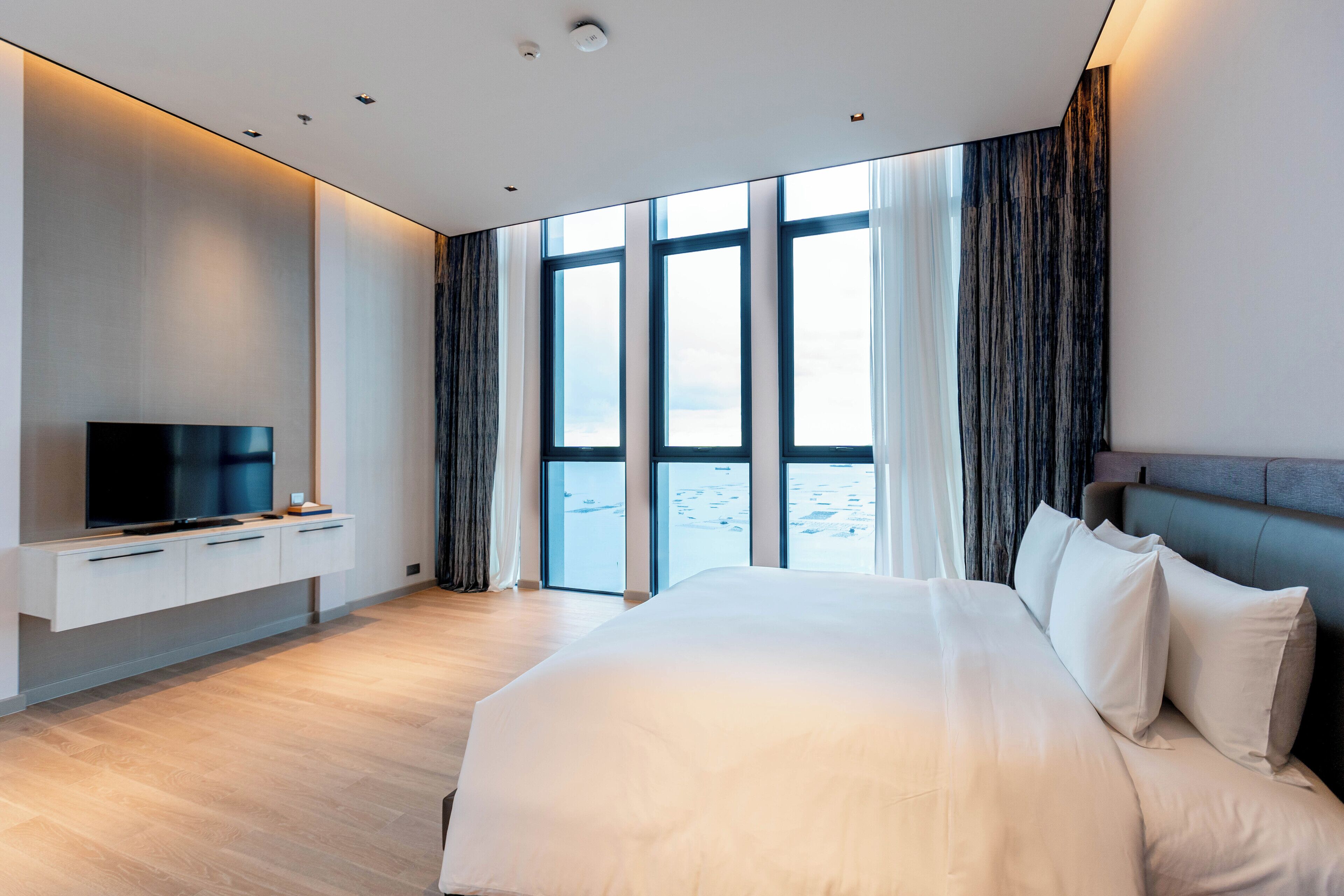 Habitación de hotel moderna con una cama grande, un televisor de pantalla plana y vistas al mar a través de amplias ventanas.