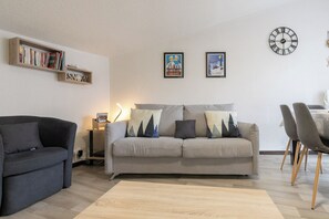 A living room with a sofa, a dark armchair, a bookshelf, a wall clock, and two framed pictures.