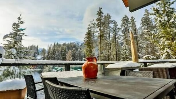 Une table avec un vase rouge, des chaises et une vue sur des arbres et des montagnes enneigés.