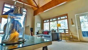 Un salon moderne avec une table basse en verre, des poutres en bois au plafond et de grandes fenêtres.